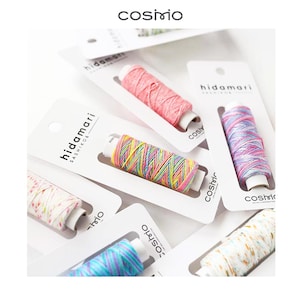 May include: Multiple spools of Hidamari Sashiko thread in various colours, including pink, multi-coloured neon, blue and purple, and white with speckles. Each spool is mounted on a white cardboard display card with the brand name Cosimo printed.