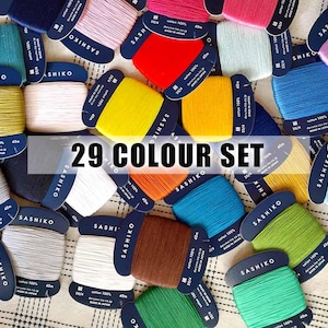 May include: A set of 29 spools of thread in various colours. Each spool is labelled "Sashiko" and has a white background with black text. The spools are arranged in a circle on a checkered fabric.