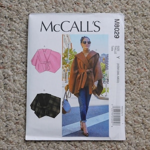 May include: McCall's M8029 sewing pattern featuring a hooded cape with a tie closure. The pattern includes size Y (XSM-SML-MED). The image shows a pink cape, a plaid cape, and a brown cape being worn.