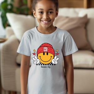 May include: A light grey t-shirt with a yellow smiley face wearing a red and white patterned hat with the letters "KC" on it. The smiley face is giving a peace sign with both hands. The text "kansas city" is printed below the smiley face.