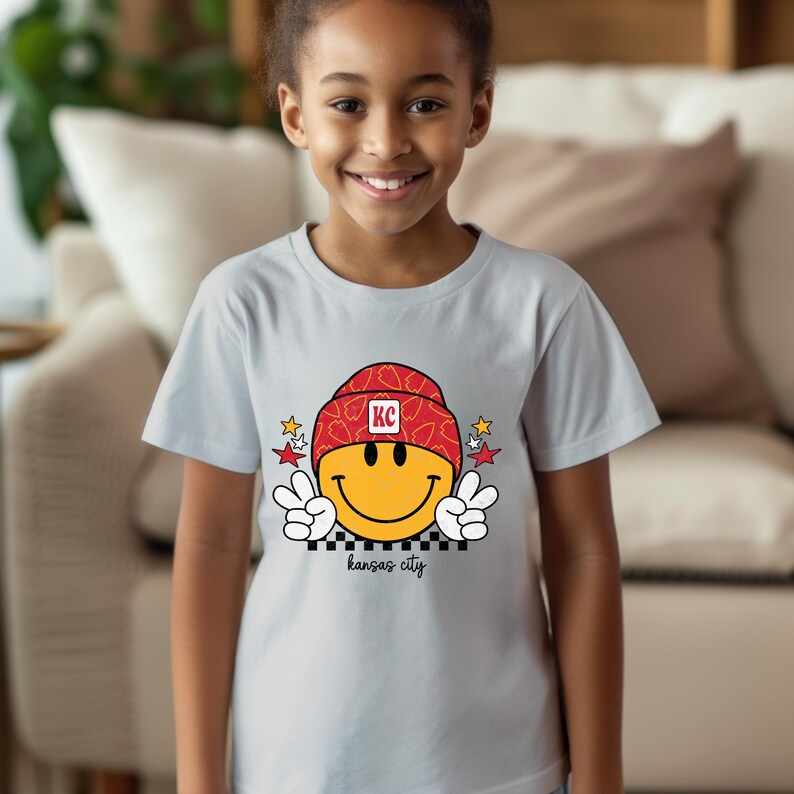 May include: A light grey t-shirt with a yellow smiley face wearing a red and white patterned hat with the letters "KC" on it. The smiley face is giving a peace sign with both hands. The text "kansas city" is printed below the smiley face.
