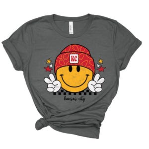 May include: A charcoal grey t-shirt with a yellow smiley face wearing a red and white hat with the letters "KC" on it. The smiley face is giving a peace sign with both hands. The text "kansas city" is printed below the smiley face.