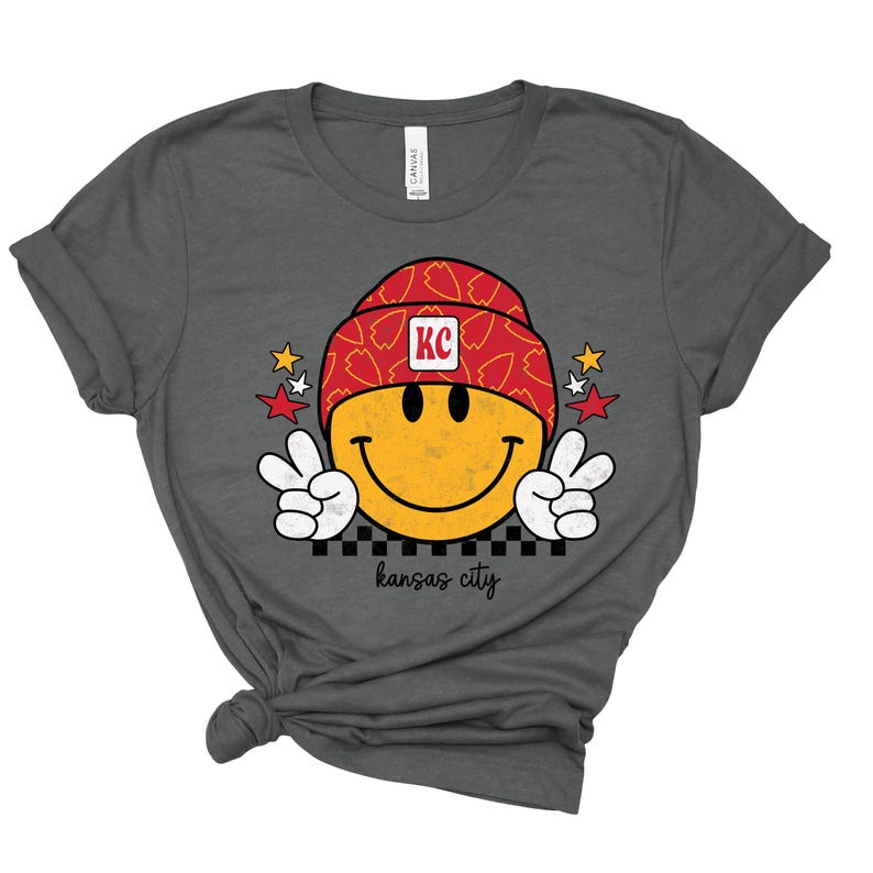 May include: A charcoal grey t-shirt with a yellow smiley face wearing a red and white hat with the letters "KC" on it. The smiley face is giving a peace sign with both hands. The text "kansas city" is printed below the smiley face.