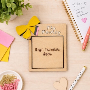 Op de afbeelding: Een rechthoekig houten bord met de tekst Mrs Mowrey en Best Teacher Ever, versierd met een gele strik in de vorm van een potlood. Het bord is gemaakt van lichtgekleurd hout met donkere gegraveerde letters en een zwarte rand rond de tekst.