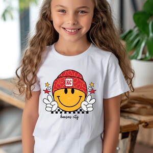 May include: White t-shirt with a yellow smiley face wearing a red and white hat with "KC" on it. The smiley face is giving a peace sign with each hand. The text "kansas city" is below the smiley face.