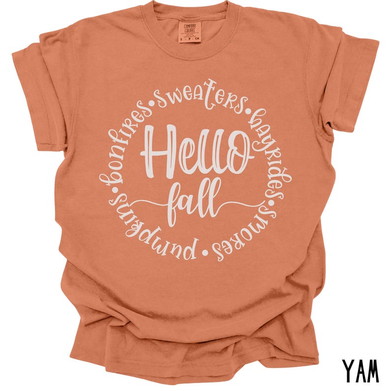 May include: A burnt orange t-shirt with a white circle graphic that says "Hello Fall" with the words "bonfires, sweaters, hayrides, s'mores, pumpkin" surrounding the text.