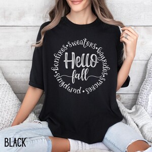 May include: Black t-shirt with a white graphic design that says "Hello Fall" surrounded by the words "bonfires, sweaters, hayrides, smokes, sundwind".