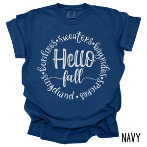 May include: A navy blue t-shirt with a white circle graphic that says "Hello Fall" surrounded by the words "bonfires, sweaters, hayrides, s'mores, pumpkin".