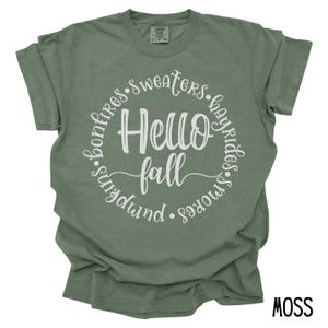 May include: A moss green t-shirt with a white circle graphic that says "Hello Fall" surrounded by the words "bonfires, sweaters, hayrides, s'mores, pumpkin".