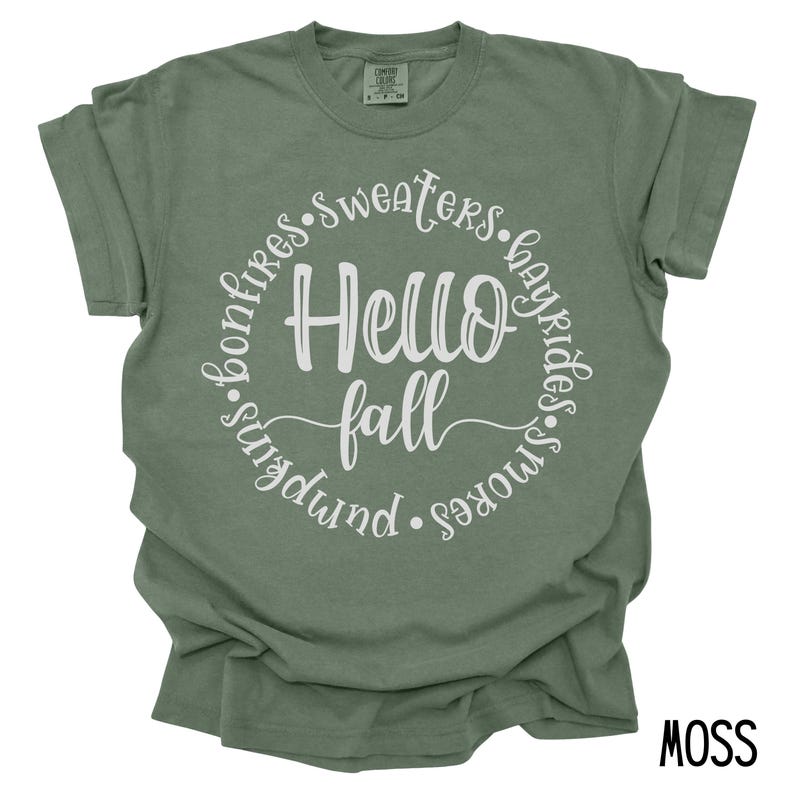 May include: A moss green t-shirt with a white circle graphic that says "Hello Fall" surrounded by the words "bonfires, sweaters, hayrides, s'mores, pumpkin".