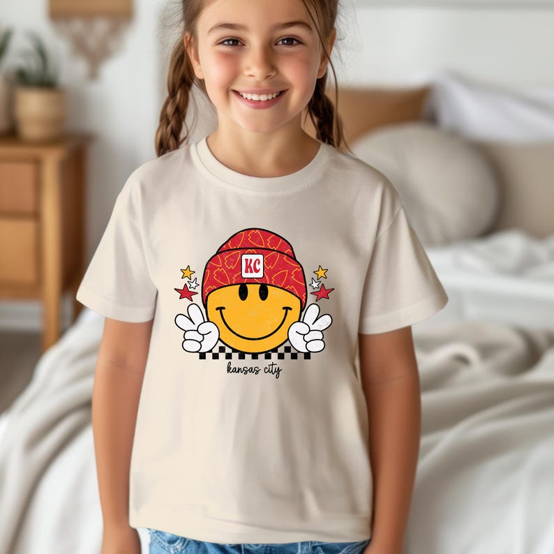 May include: A white t-shirt with a yellow smiley face wearing a red and white patterned hat with the letters "KC" on it. The smiley face is giving a peace sign with both hands. The text "kansas city" is printed below the smiley face.