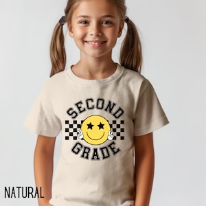May include: A beige t-shirt with the word "NATURAL" at the bottom. The shirt features a graphic with a yellow smiley face with star eyes and peace signs, and the words "SECOND GRADE" in black.