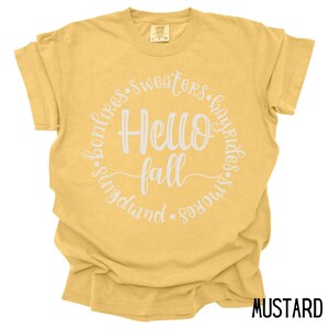 May include: A mustard yellow t-shirt with a white circle graphic that says "Hello Fall" and is surrounded by the words "bonfires, sweaters, hayrides, s'mores, pumpkin".