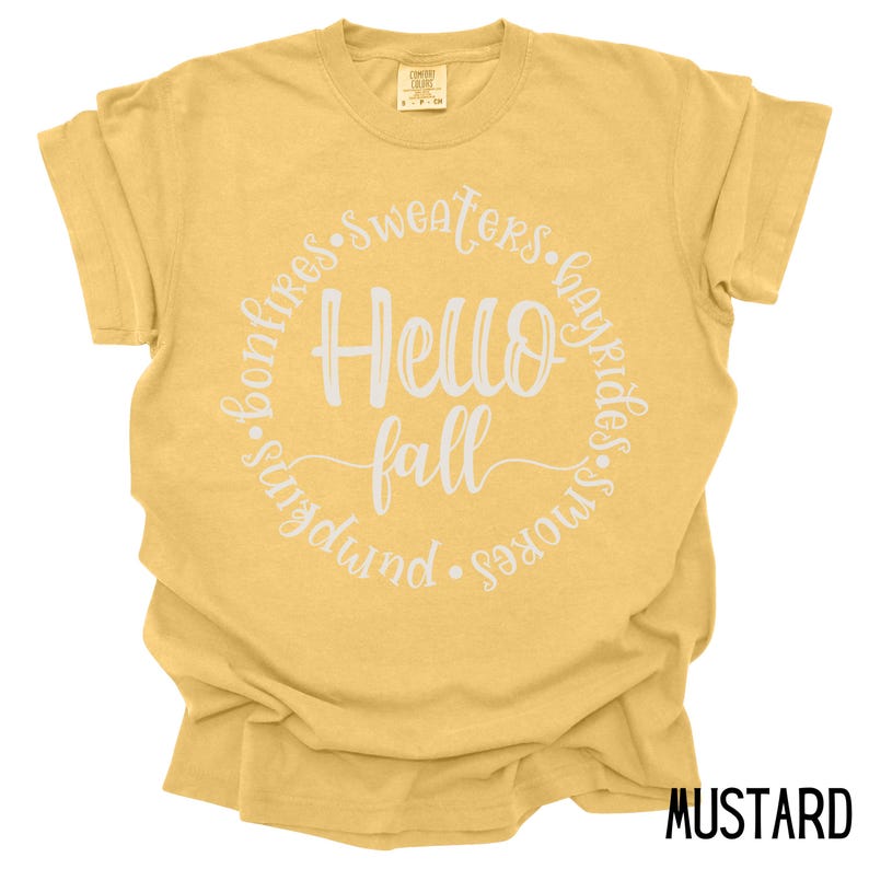 May include: A mustard yellow t-shirt with a white circle graphic that says "Hello Fall" and is surrounded by the words "bonfires, sweaters, hayrides, s'mores, pumpkin".