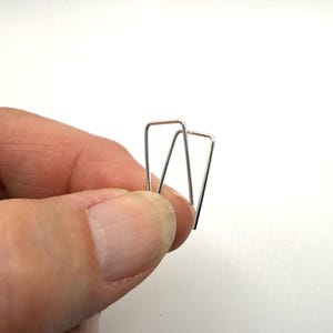 May include: A pair of small, silver-colored metal earrings. Each earring has a rectangular top with a V-shaped bottom, creating a geometric design. The earrings are held against a white background.