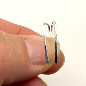 May include: A pair of silver-colored ear cuffs. The ear cuffs are held between fingers and thumb. The ear cuffs have a simple, minimalist design, with a curved top and straight, flat sides. The metal has a slightly hammered texture.