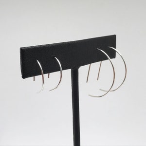 May include: Three silver hoop earrings of varying sizes displayed on a black earring stand.