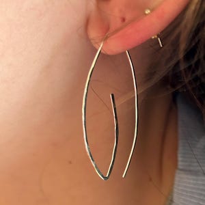 May include: A pair of silver hoop earrings with a unique, slightly asymmetrical design. The earrings are thin and delicate, and they hang from the earlobe.