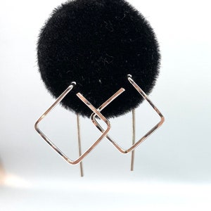May include: A pair of silver square hoop earrings with a simple design. The earrings are hanging from a black velvet display stand.