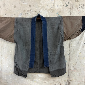 May include: A vintage-style kimono jacket in shades of brown, gray, and navy blue. The jacket features a corduroy-like texture and a contrasting navy blue collar and trim. The sleeves are wide and the jacket is hanging on a wooden rod.