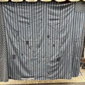 Könnte beinhalten: Ein großes, vertikal gestreiftes Textil in Grau- und Blautönen mit einem Patchwork-Design. Das Textil hängt, möglicherweise als Vorhang oder Wandbehang. Die Ränder weisen ein kontrastierendes Muster aus weißen Punkten auf schwarzem Hintergrund auf.