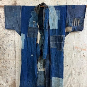 May include: A vintage indigo blue jacket with patchwork details. The jacket features various shades of blue fabric, with visible stitching and a worn aesthetic. The garment is displayed hanging, showcasing its full length and design.