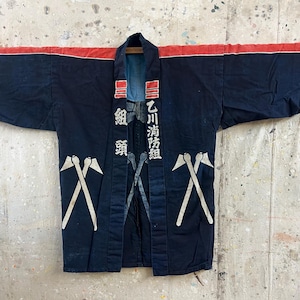 Könnte beinhalten: Eine dunkelblaue japanische Hanten-Jacke mit einem roten horizontalen Streifen über den Schultern, weißen japanischen Kanji-Schriftzeichen auf den Vorderteilen und weißen grafischen Drucken von gekreuzten Feuerwehraxten.
