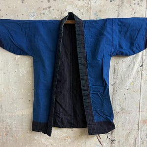 Może przedstawiać: Niebiesko-czarna, zabytkowa japońska kurtka kimono. Kurtka jest otwarta, odsłaniając czarne, prążkowane wnętrze. Rękawy są szerokie, a kurtka wykonana jest z teksturowanej tkaniny. Kurtka jest prezentowana na drewnianym pręcie.