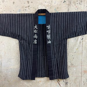 Puede incluir: Una chaqueta happi de rayas negras y grises con mangas anchas y un cuello negro sólido. El cuello muestra caracteres kanji japoneses blancos que dicen 大松商店 a la izquierda y 味噌醤油 a la derecha. El forro interior es de tela azul brillante.