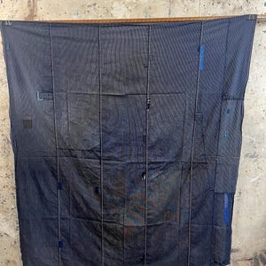May include: A large, dark blue textile with visible stitching and patches. The fabric is hung from a wooden rod, displaying a patchwork design with vertical and horizontal seams. Patches of blue fabric are sewn onto the main textile, adding visual interest.