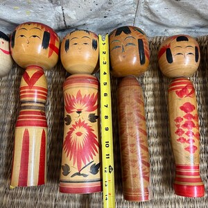 May include: A collection of wooden Kokeshi dolls, each with a round head and cylindrical body. The dolls are painted with facial features, and some have decorative patterns in red and black. The tallest doll measures approximately 33 cm.