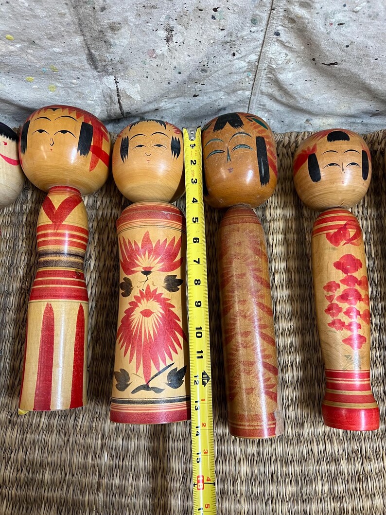 May include: A collection of wooden Kokeshi dolls, each with a round head and cylindrical body. The dolls are painted with facial features, and some have decorative patterns in red and black. The tallest doll measures approximately 33 cm.