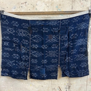 May include: A rectangular indigo blue textile featuring a white ikat-style diamond pattern. The fabric displays visible mending patches in darker blue tones and is finished with a light-coloured fabric border along the top edge with ties.