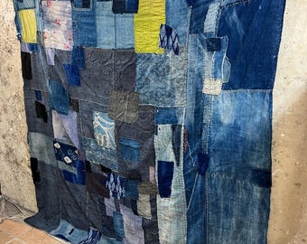 May include: A large patchwork quilt made from various shades of blue denim fabric. The quilt is composed of many different sized squares and rectangles, and it has been stitched together with a variety of different coloured threads. The quilt is hanging on a wall, and it is lit by natural light.
