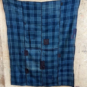 Puede incluir: Una pieza textil rectangular con un patrón de cuadros en azul y azul oscuro. La tela está construida con múltiples paneles unidos por costuras visibles e incluye varios parches cuadrados pequeños de color azul oscuro cosidos.