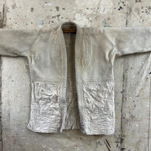 Peut inclure: Un uniforme d'arts martiaux blanc cassé, probablement un judogi, est présenté. La veste présente un tissage texturé, des coutures renforcées et des patchs sur le bas du devant. La veste ouverte est suspendue à une tringle en bois sur un fond moucheté.