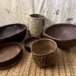 May include: A collection of handcrafted, rustic bowls and containers. The bowls vary in size and shape, with dark brown and tan tones. One tall, cylindrical container and several oval and round bowls are visible, all with a natural, aged appearance.