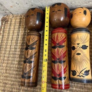 May include: Three wooden Japanese Kokeshi dolls. The tallest doll is approximately 48 cm tall. The dolls have painted faces and decorative patterns in black and red. The dolls are displayed on a woven mat.