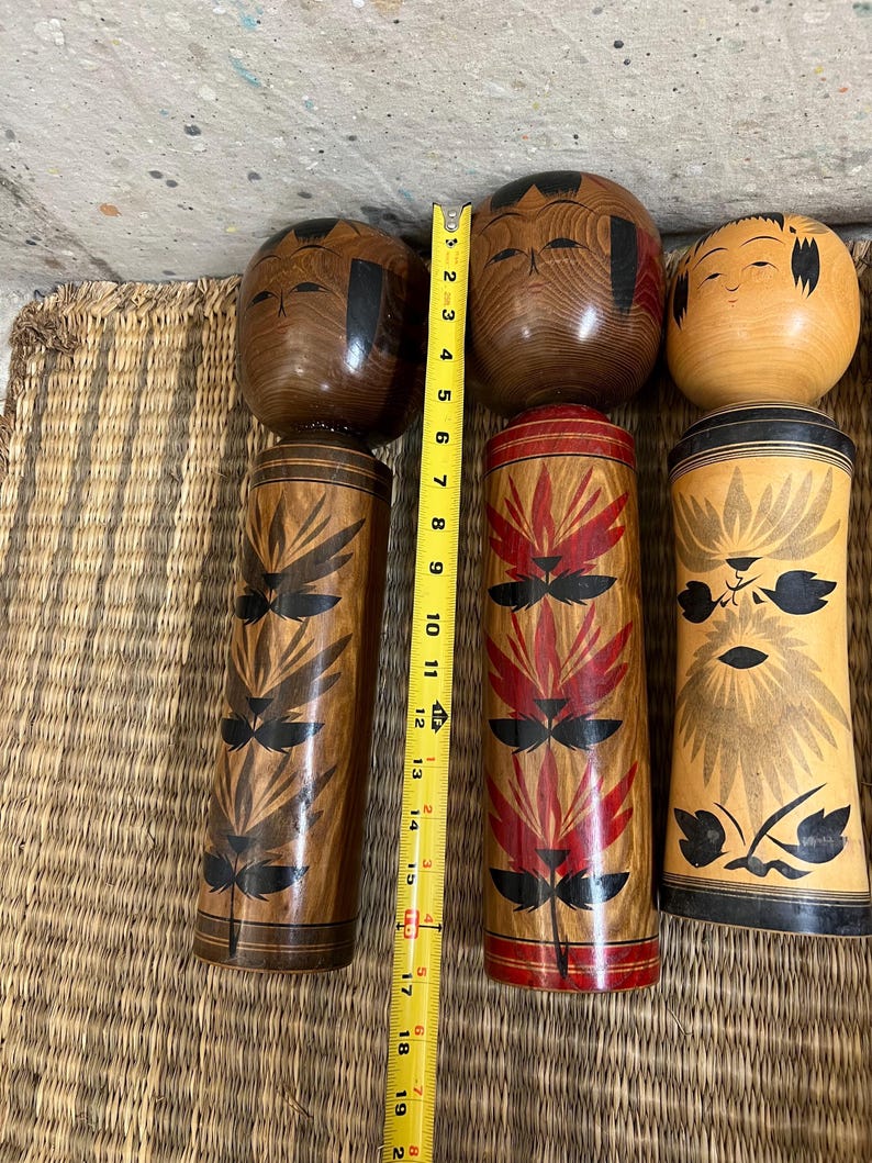 May include: Three wooden Japanese Kokeshi dolls. The tallest doll is approximately 48 cm tall. The dolls have painted faces and decorative patterns in black and red. The dolls are displayed on a woven mat.