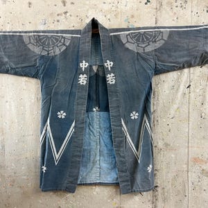 May include: A faded blue cotton jacket featuring white Japanese kanji characters on the front lapels, circular crest patterns on the shoulders, and white cherry blossom motifs with geometric zigzag lines along the lower hem and sleeve edges.