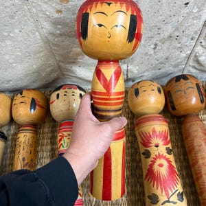 May include: A selection of traditional Japanese Kokeshi dolls. The wooden dolls have painted details, including facial features and decorative patterns. The dolls are mainly in shades of brown, red, and yellow.