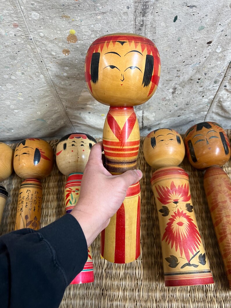 May include: A selection of traditional Japanese Kokeshi dolls. The wooden dolls have painted details, including facial features and decorative patterns. The dolls are mainly in shades of brown, red, and yellow.