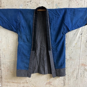 Puede incluir: Una chaqueta azul y negra estilo kimono. La chaqueta tiene un cuerpo azul oscuro con ribetes negros en las mangas y los bordes. El interior es gris oscuro con rayas verticales. La chaqueta está colgada de una barra de madera.