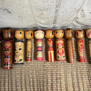 May include: A selection of twelve Japanese Kokeshi dolls. The dolls feature wooden cylindrical bodies and rounded heads with painted faces. The bodies are adorned with diverse patterns, including floral designs, stripes, and geometric shapes. The dolls vary in size and colour.