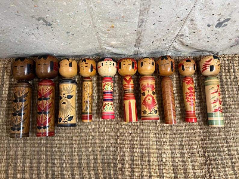 May include: A selection of twelve Japanese Kokeshi dolls. The dolls feature wooden cylindrical bodies and rounded heads with painted faces. The bodies are adorned with diverse patterns, including floral designs, stripes, and geometric shapes. The dolls vary in size and colour.