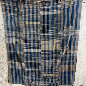 May include: A rectangular textile featuring a patchwork design of blue, tan, and off-white plaid patterns. The fabric is composed of multiple vertical panels stitched together, displaying visible mending, frayed edges, and varied weave densities.