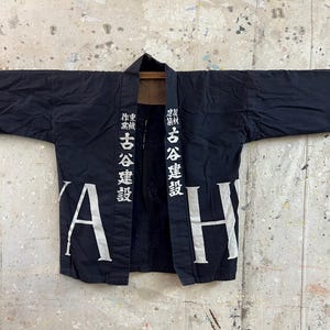 Peut inclure: Veste de travail japonaise noire avec texte japonais blanc. La veste est ouverte, révélant un intérieur marron. Les manches sont étendues, et la veste est suspendue à une tige en bois sur un fond moucheté.
