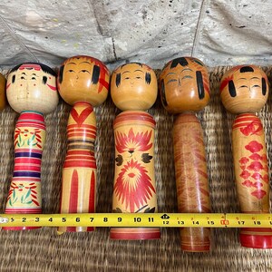 May include: Six traditional Japanese Kokeshi dolls with wooden bodies and heads. The dolls have painted faces and decorative patterns in red, yellow, and black. A measuring tape indicates the dolls are approximately 15 cm to 18 cm in height.