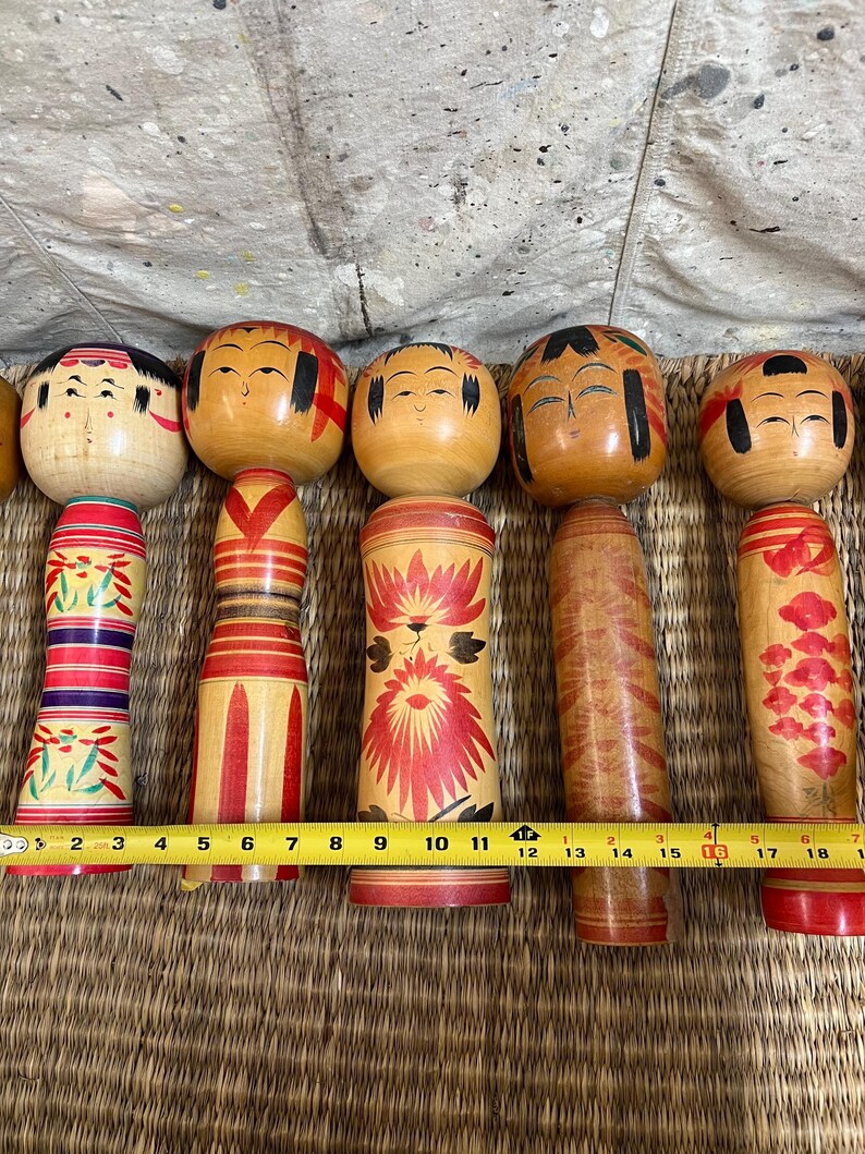 May include: Six traditional Japanese Kokeshi dolls with wooden bodies and heads. The dolls have painted faces and decorative patterns in red, yellow, and black. A measuring tape indicates the dolls are approximately 15 cm to 18 cm in height.
