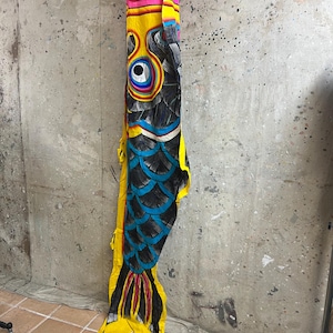 May include: A vibrant carp streamer, or koinobori, is suspended against a textured wall. The fish-shaped decoration has a yellow body, black scales, and a red top. The design incorporates blue, black, and yellow details, with a yellow tail.